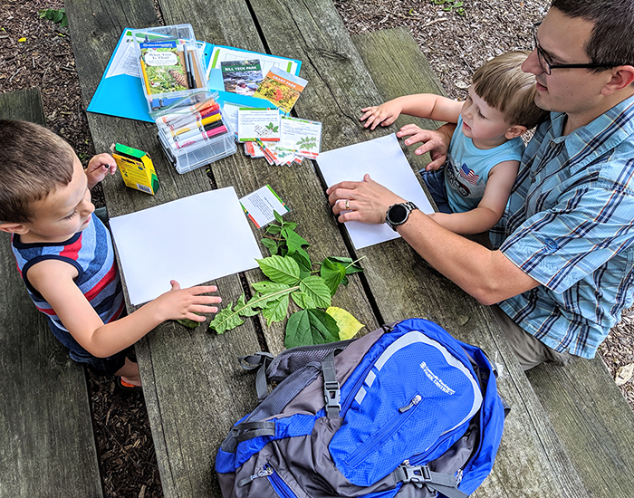 father and two children leaf rubbing at picnic table with adventure backpack