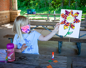 girl in mask sitting at picnic table holding up flower artwork