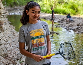 teenage girl standing in Holes Creek with a net. Wearing a CWPD summer camp t-shirt.