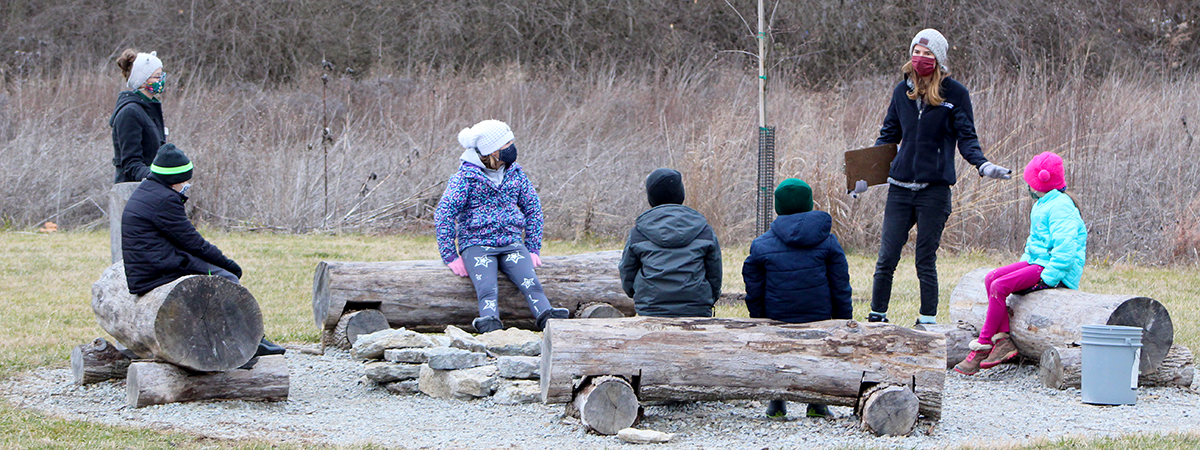 Five children sitting around a fire pit on logs