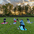 five people doing yoga on the lawn with the sunset behind pine trees