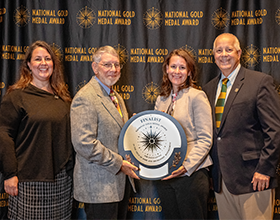 four people holding a gold medal plaque in front of a photo backdrop