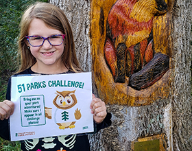 child holding a 51 Parks Challenge sign in front of a tree carving of a fox