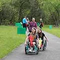 six people on a paved path through a grassy area, two people are using wheelchairs