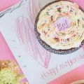 a mothers day cake placed on top of a handmade mothers day card on a pink background